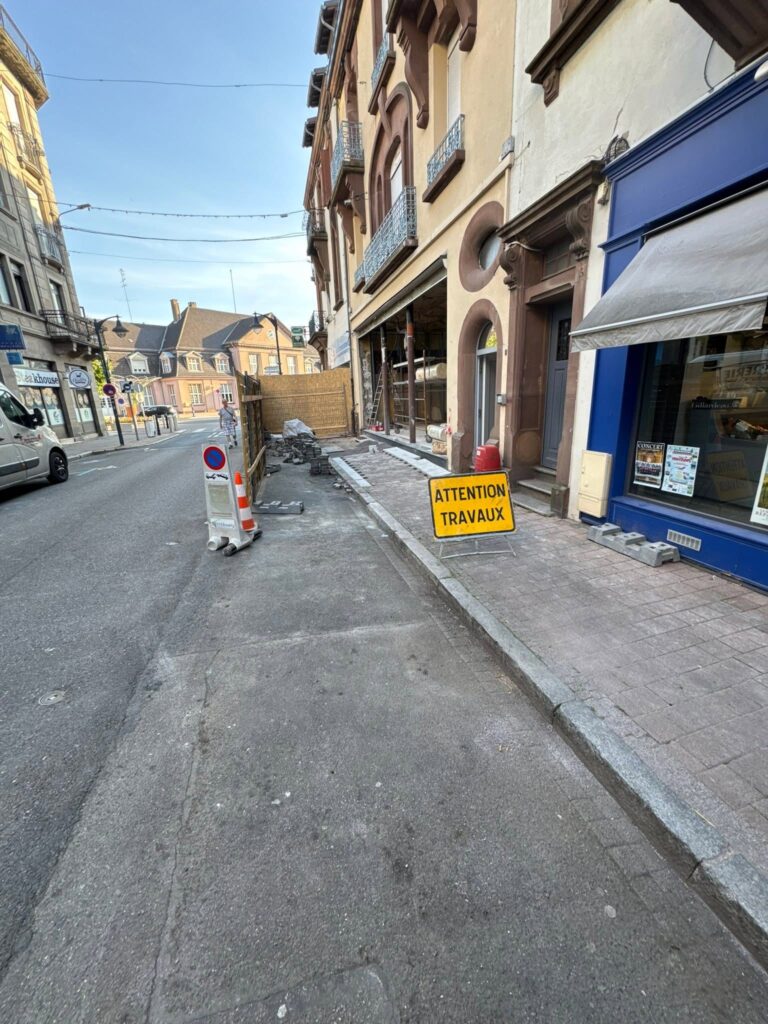 Street under construction: sidewalk blocked on the right with a yellow 'ATTENTION TRAVAUX' sign and rubble.