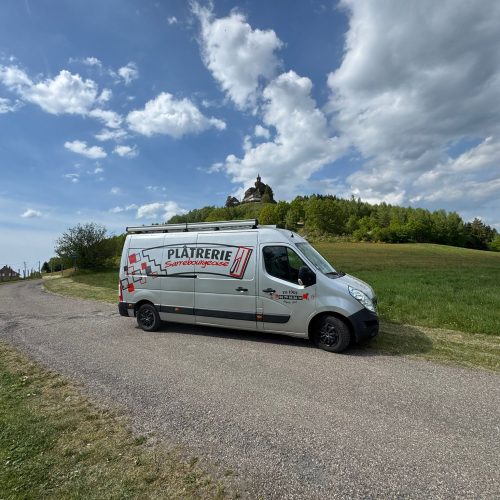 Silver branded van parked on a gravel rural road with grassy field, hilltop house and trees in the distance under a blue sky with clouds.